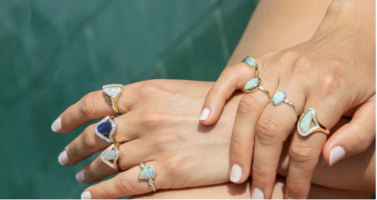 A woman's hands, heavily decorated with opal rings, photographed against a backdrop of turquoise water